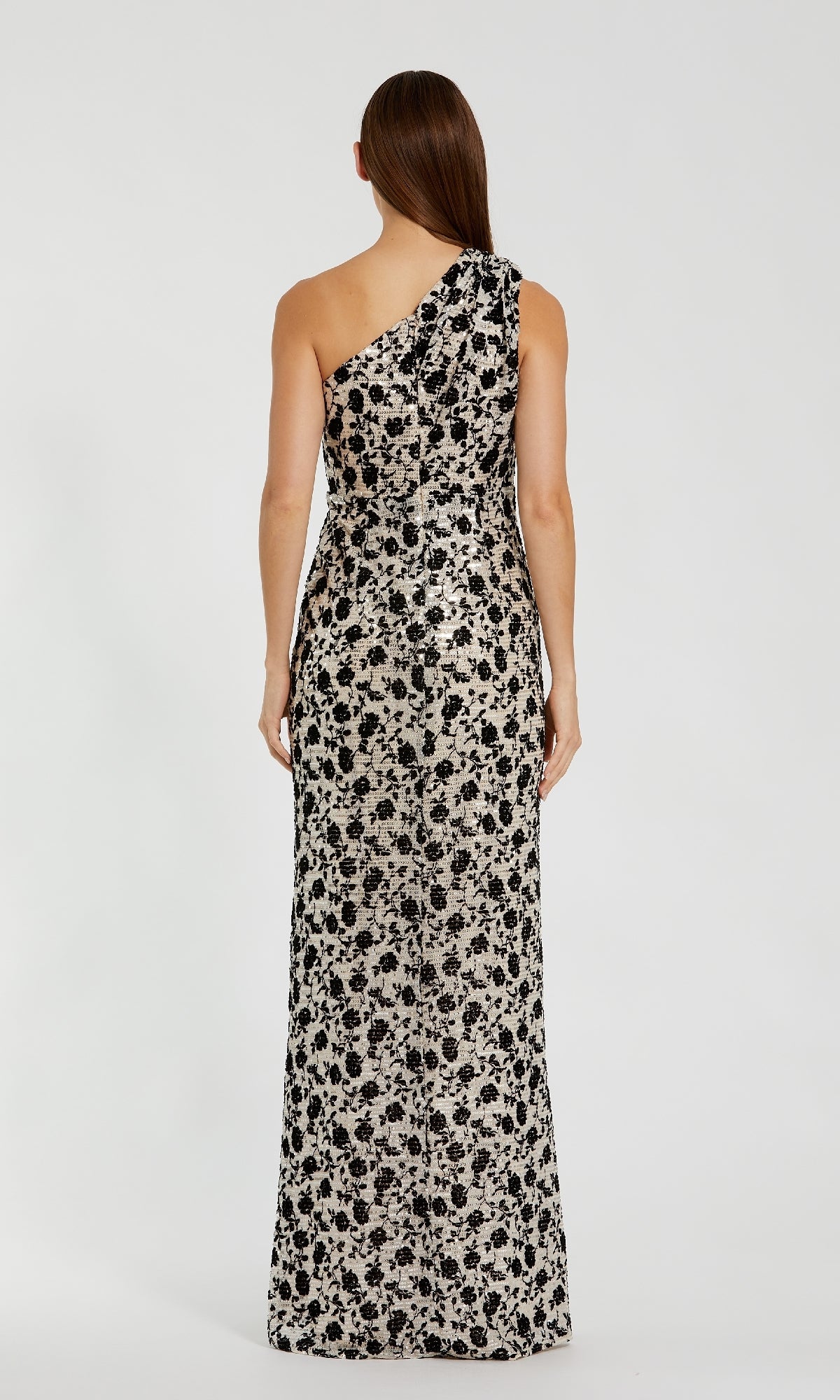 A woman faces away, wearing the Mac Duggal 27626 One-Shoulder Sequin Formal Dress— a floor-length champagne gown featuring black floral patterns on a light background.